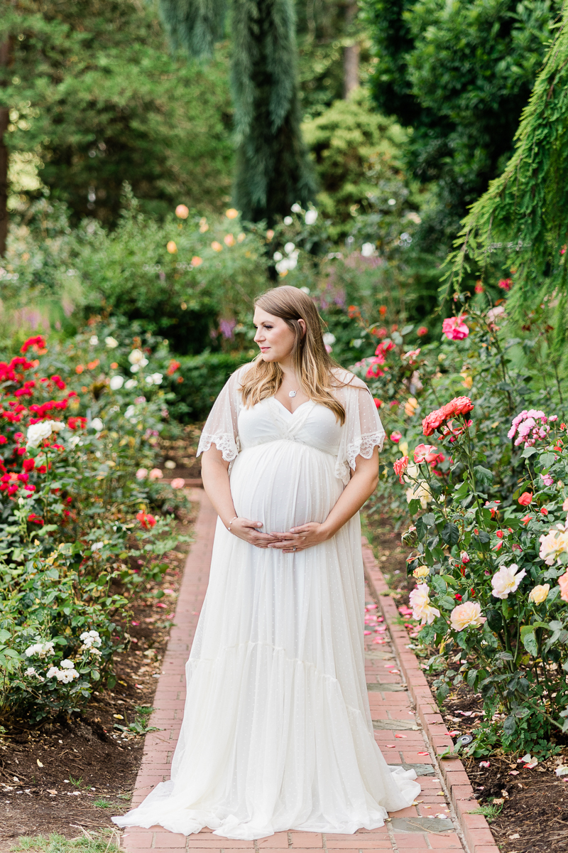 pregnant mom in white gown holding her bump in a garden and looking off into the distance unfurling birth