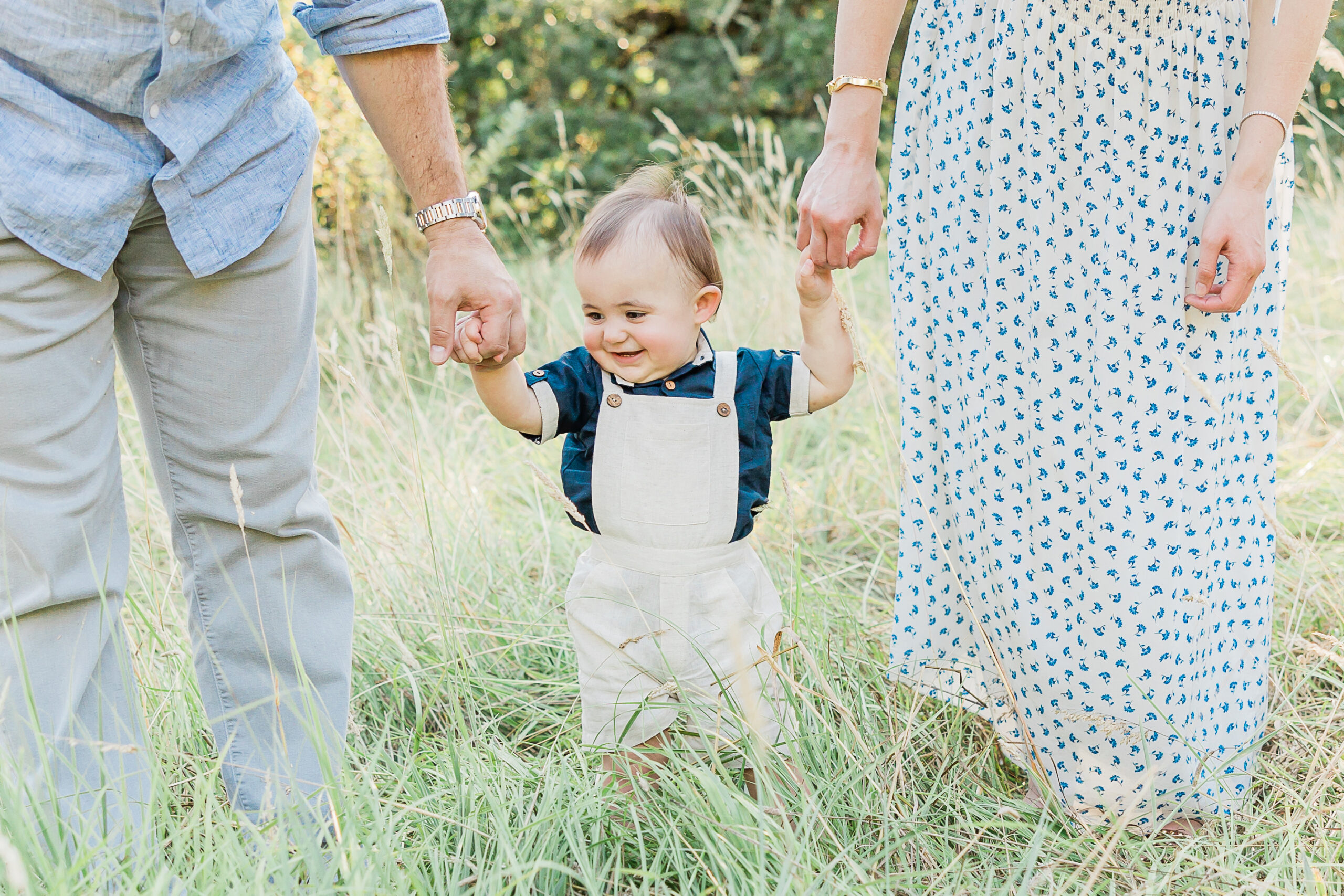 Little boy in tan overalls standing between his mom and dad in a field Hanna Andersson Portland