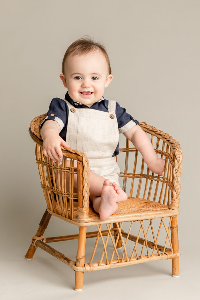 little boy in tan overalls and blue shirt sitting in a wicker chair