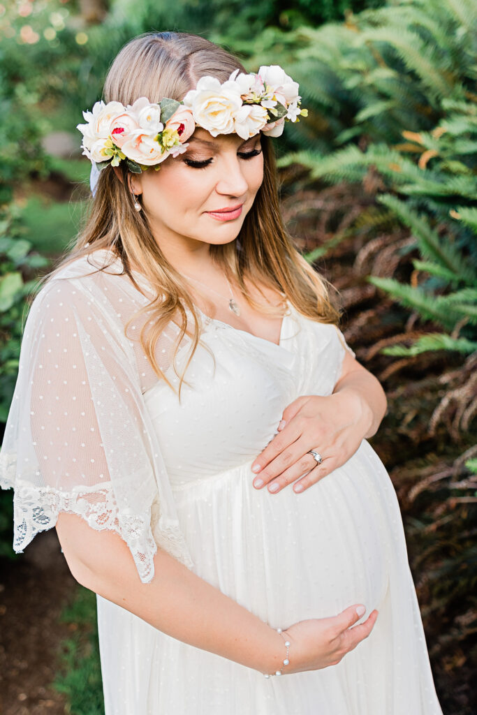 mom to be in white maternity gown holding her bump and wearing a flower crown in a garden Splendid Portland
