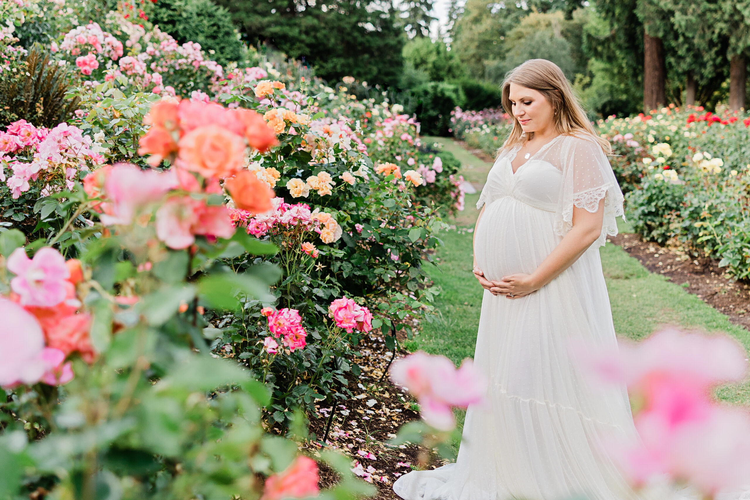 pregnant woman in long white lace maternity gown holding her bump in a garden of pink flowers Splendid Portland