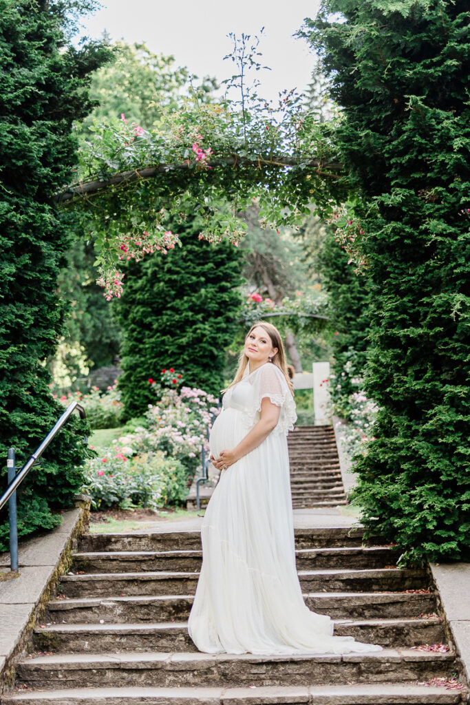 mom to be in white maternity gown standing on stairs in front of a large hedge arch
