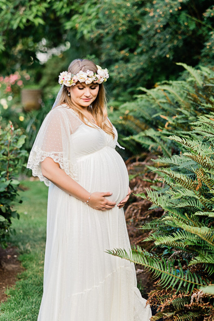 pregnant woman in white gown holding her bump and wearing a flower crown in a garden Hanau Doula