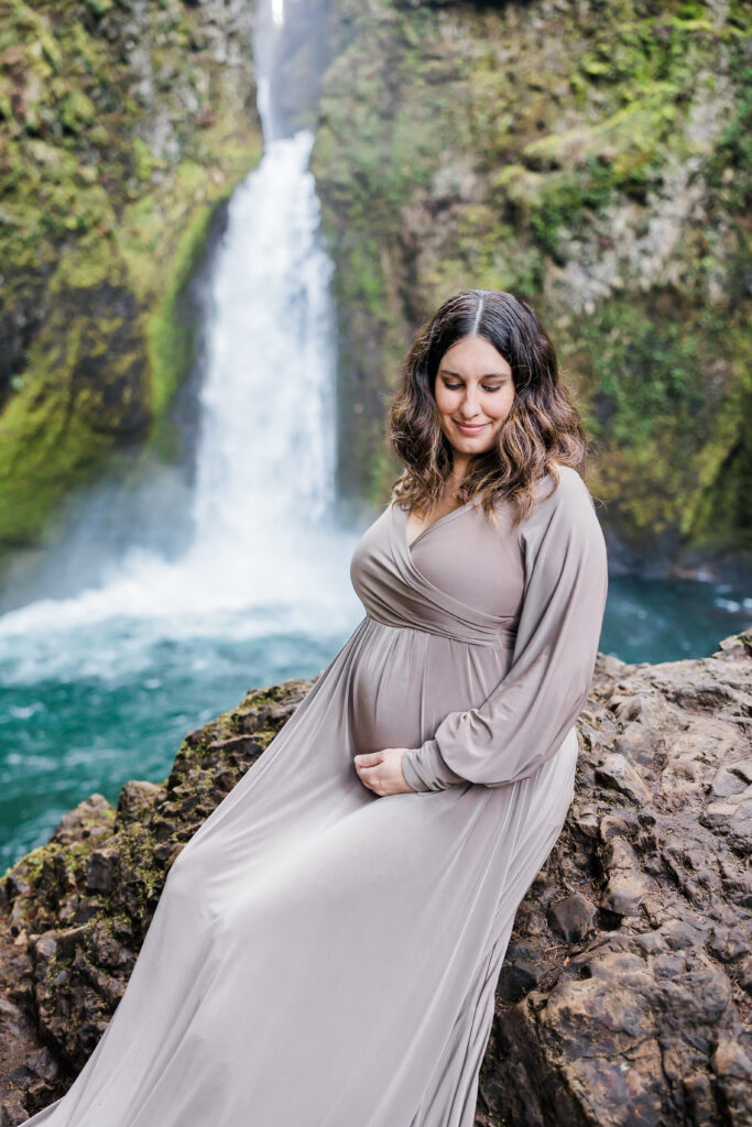 mom to be in grey maternity gown sitting on a rock in front of a waterfall near Portland Oregon