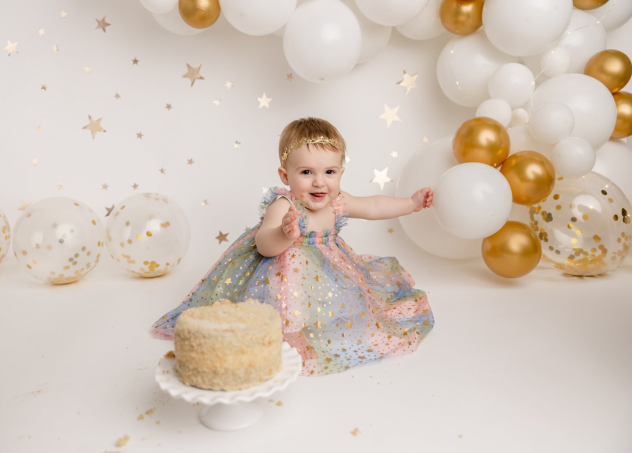little girl in a rainbow dress sitting on a floor with a cake and gold and white balloons