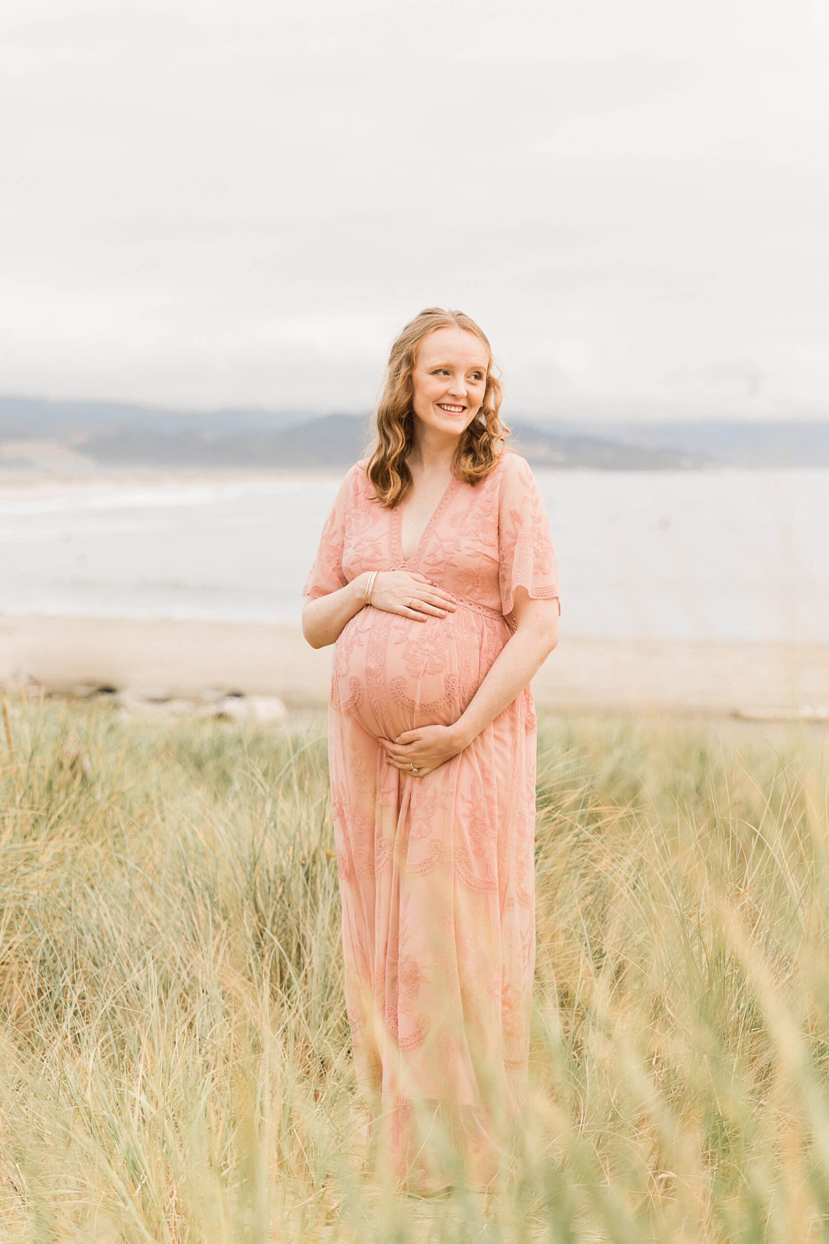 mom to be in pink maternity gown in a field of grass near the water in Portland birthing stone doula
