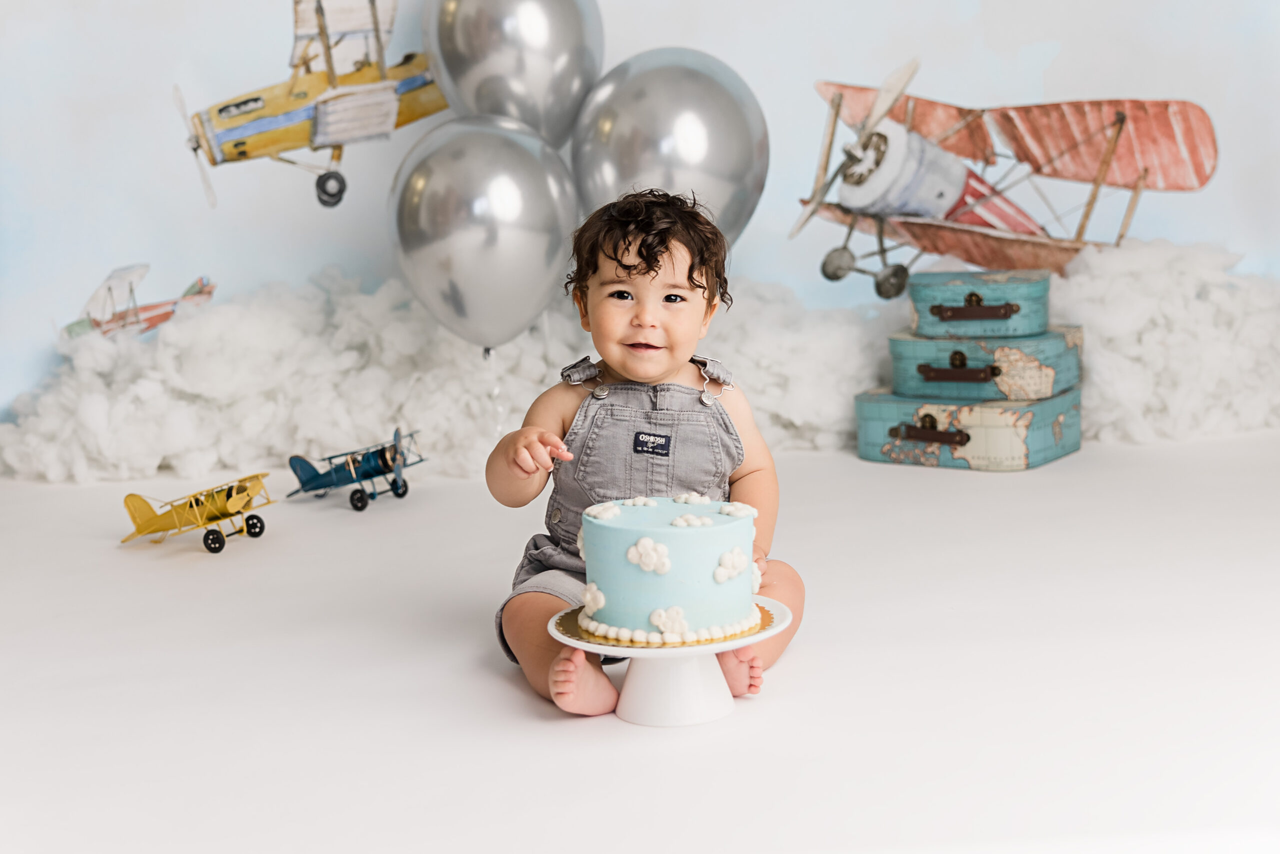 little boy celebrating his first birthday in grey overalls with airplanes behind him cuddlebugzz